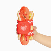 Jouet de fouille Le Hot Dog en peluche avec son pain, sa moutarde et son ketchup, tenu à la main
